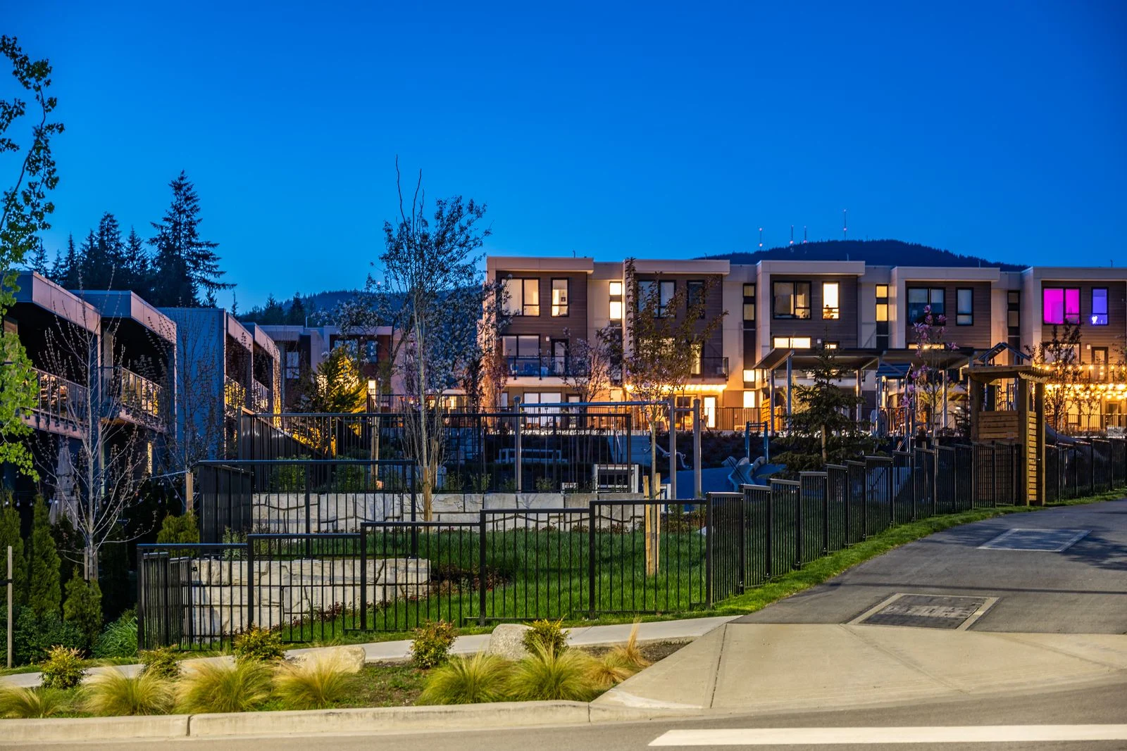 Lit townhomes and fenced park planting at dusk at Seymour Village Amenity Park