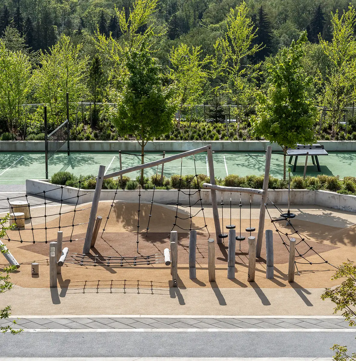 Timber play structure with rope net on a sand surface, surrounded by trees
